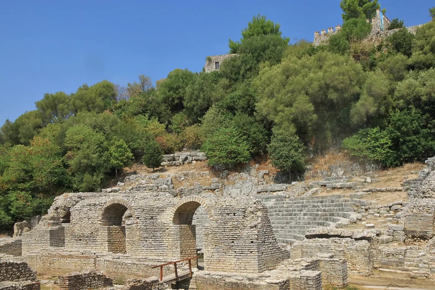 Stone seating and arched passages of Butrint's ancient theatre below a wooded hill crowned by fortress walls