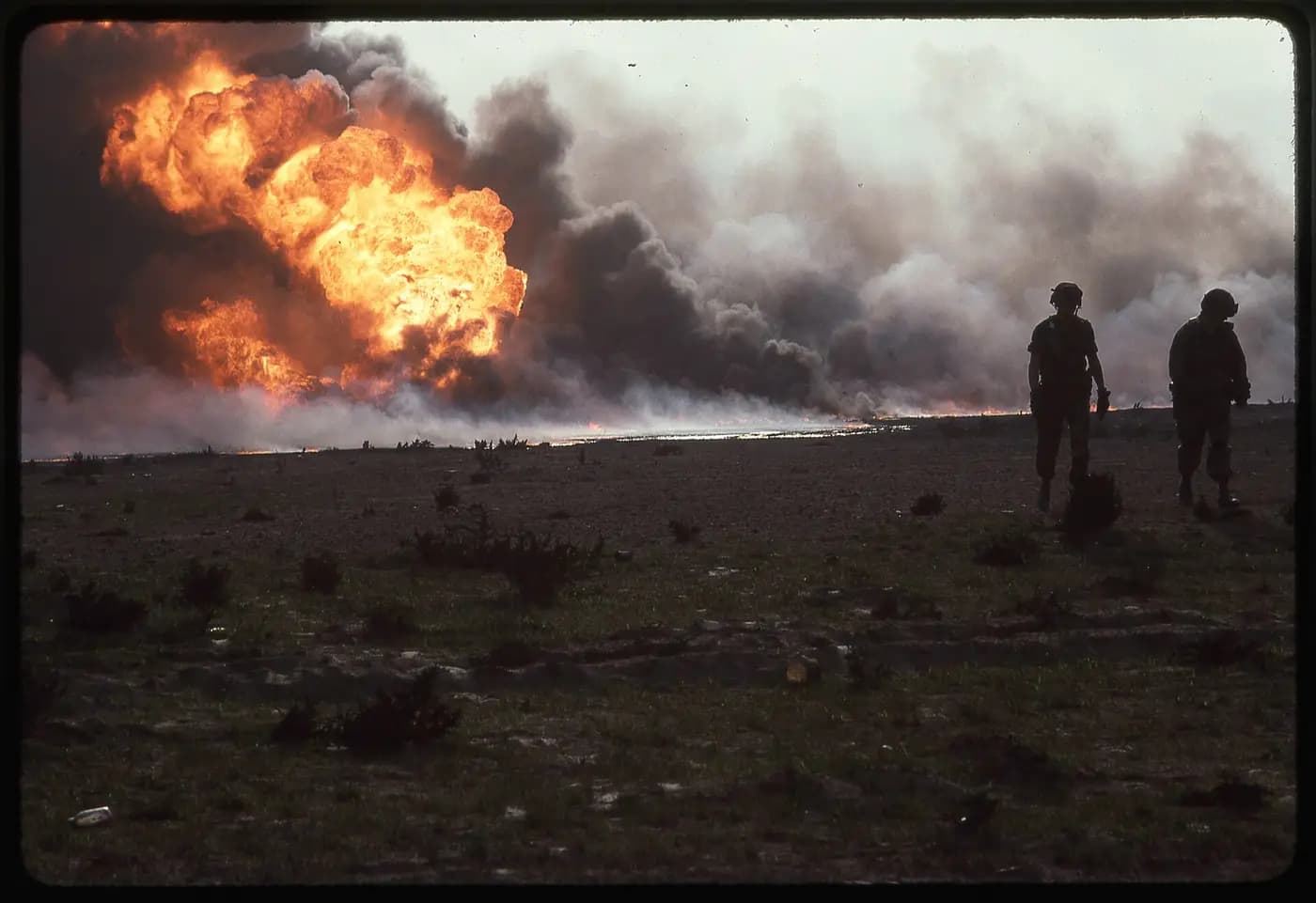 A Kuwaiti oil well burning in the desert in March 1991, with two figures walking nearby